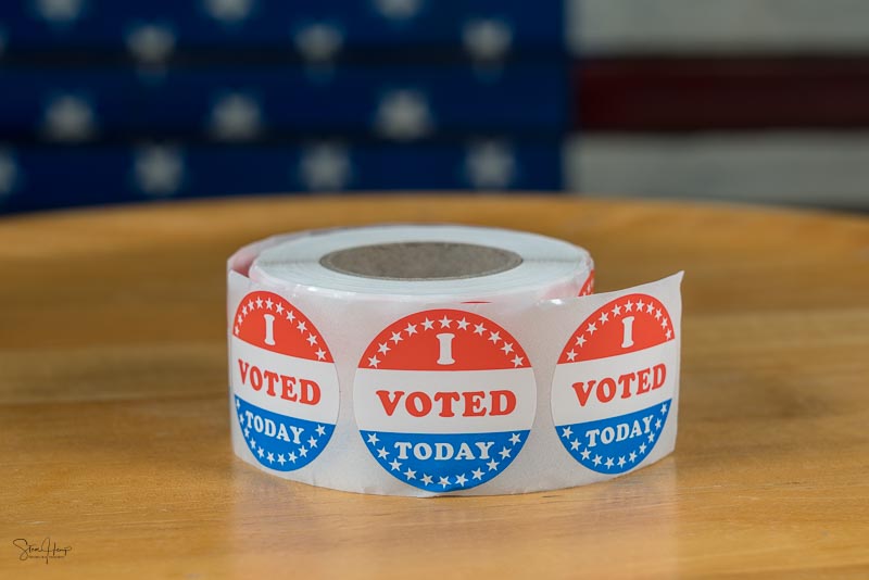 Roll of I Voted Today paper stickers on table with US Flag in ba